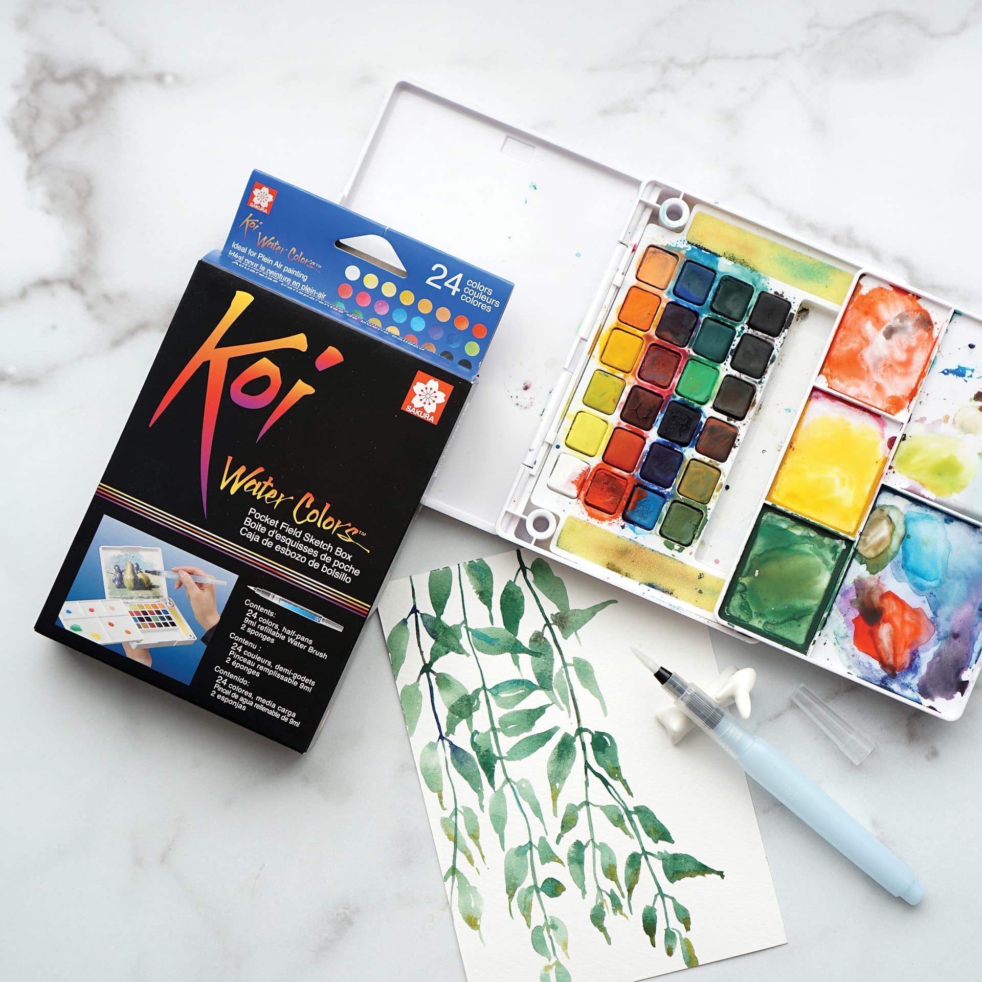 Sakura Koi Watercolor Half Pan - Set of 24 Sketch Box, Assorted Colors box beside an open set and a painting of leaves
