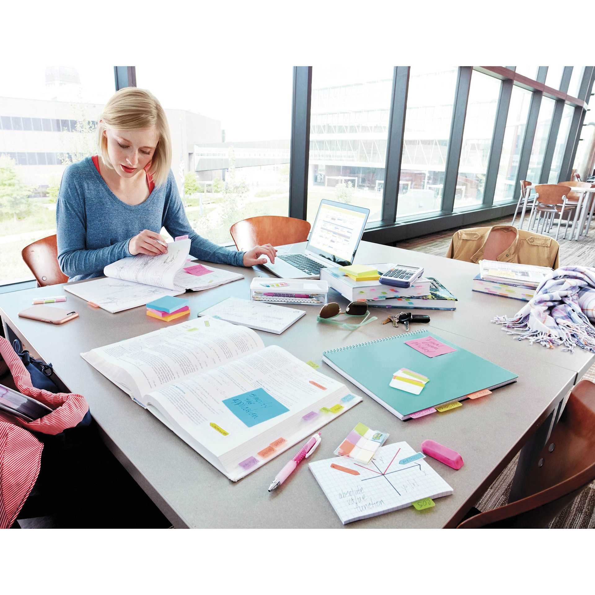 Post-it Page Markers marking pages in large book and holding notes on various office supplies sitting atop a table