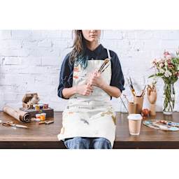 Wear'm White Apron - Artist sitting on table in paint-spattered apron with art supplies around her